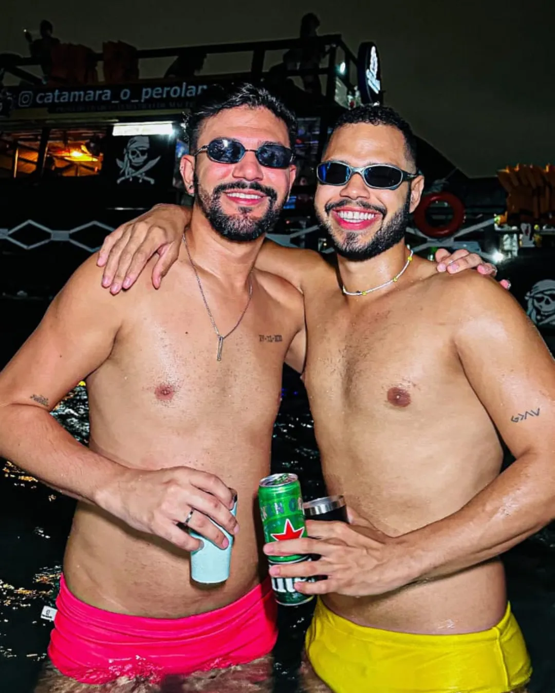 Dois amigos na água, bebida na mão, o catamarã atrás